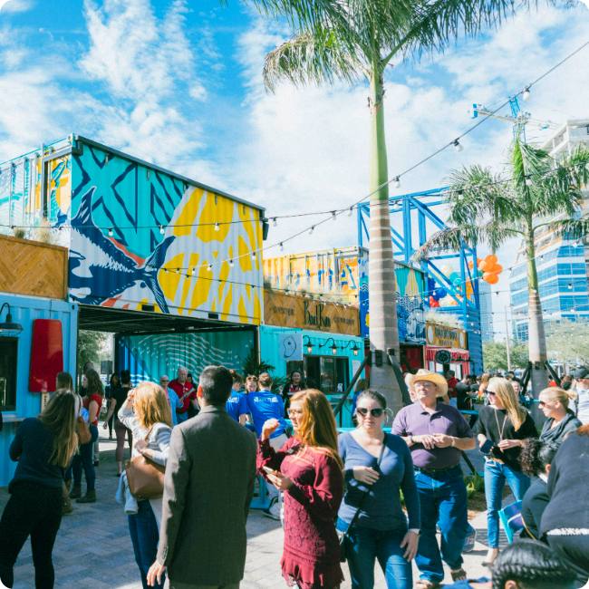 a crowd of people gathers in front of shipping containers housing restaurant stalls at sparkman wharf in tampa